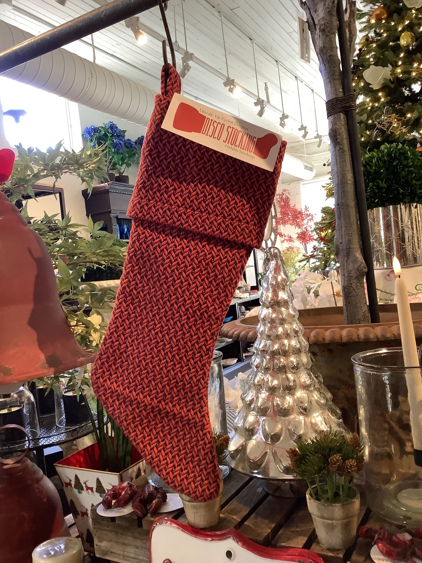 In a shop adorned with festive decorations such as a silver tree, candles, and greenery, a red knitted Christmas stocking made from recycled fabric is showcased. A label reveals the name: Disco Memo Stocking.