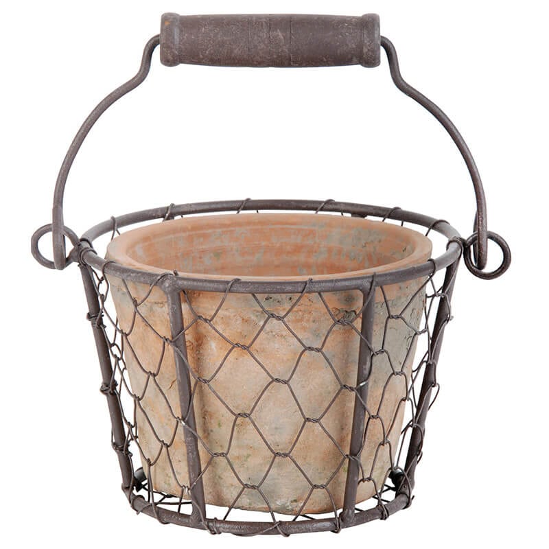 The Aged Terracotta Flower Pot in Metal Basket w/Handle combines a handcrafted rustic terra cotta pot with a metal wire basket, complete with a woven design and a wooden grip on the handle, offering an indoor/outdoor decoration that exudes a charming vintage appeal.