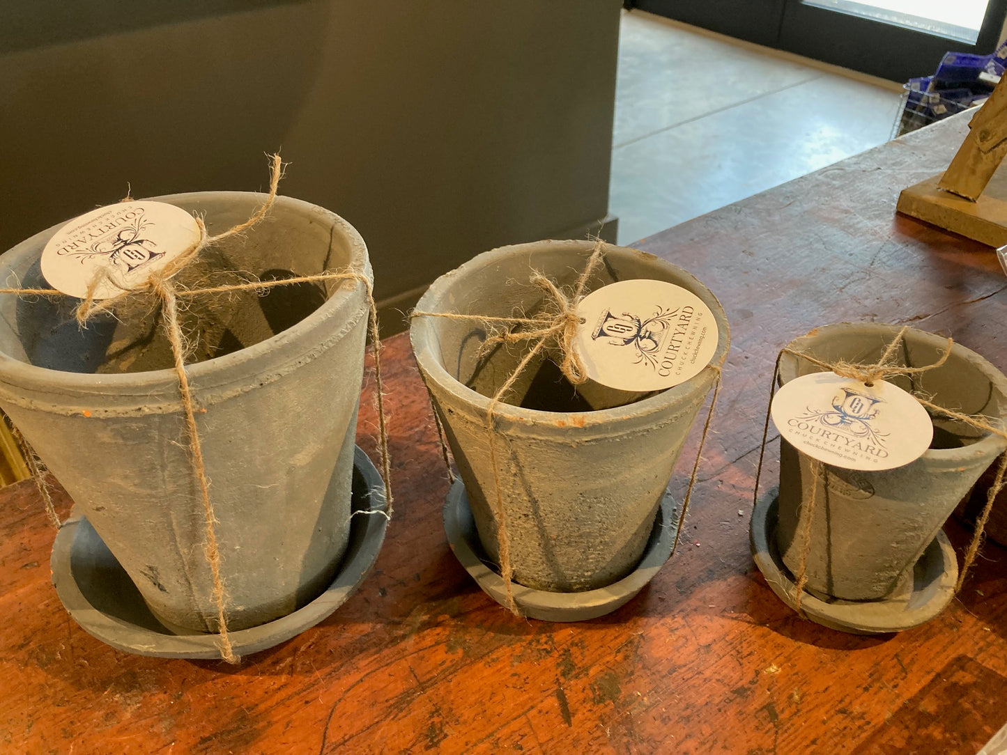 Three small aged terracotta round flower pots, each paired with a matching saucer, are elegantly arranged on a wooden table. Each pot is adorned with twine and features an attached round tag. This charming rustic set, ideal for a Tuscan-themed patio, is complemented by the backdrop of a gray wall with glass doors.